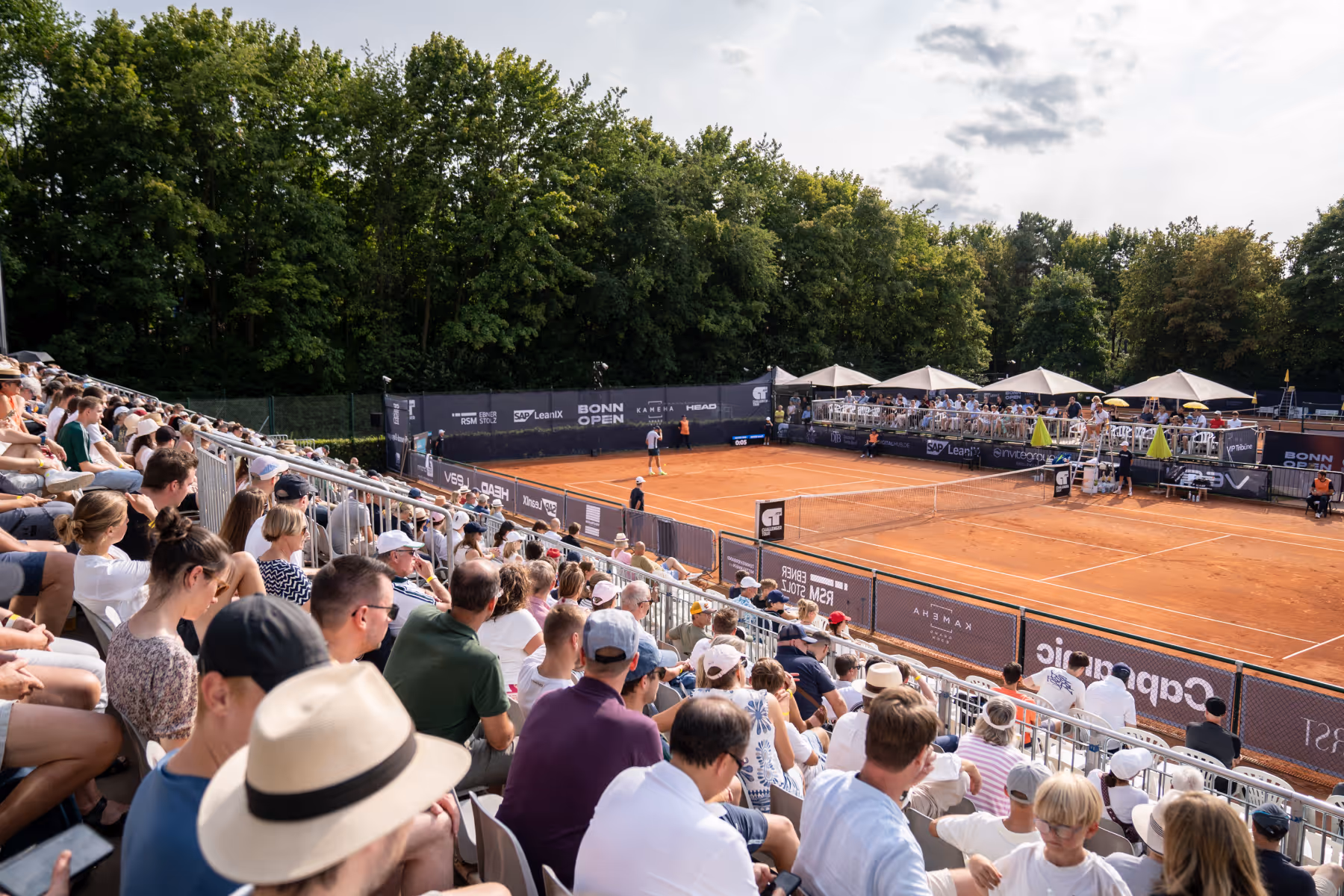 Zuschauer sitzen auf Tribünen und verfolgen ein Tennismatch auf einem Sandplatz im Freien bei sonnigem Wetter.