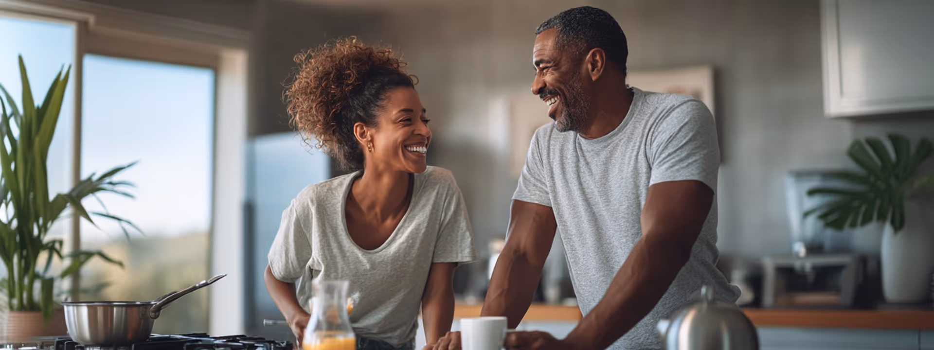 Smiling couple in casual clothes leaning on a kitchen counter with a pot on the stove and plants in the background.