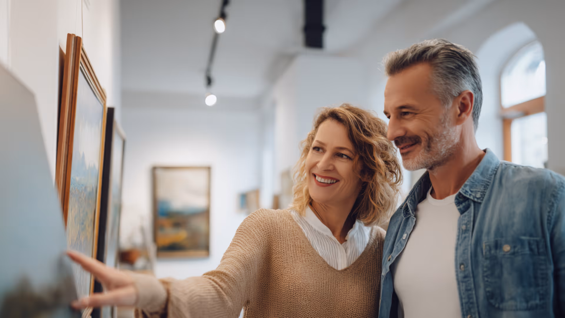 Smiling middle-aged couple looking at and discussing framed artwork in a bright gallery.