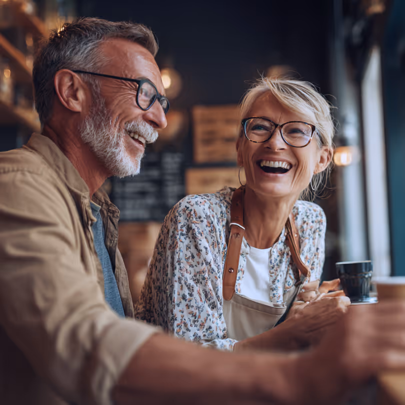 Smiling middle-aged man and woman wearing glasses sitting together at a coffee shop counter.