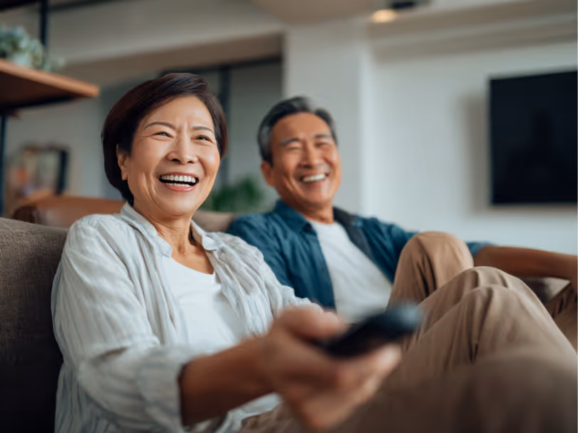Smiling elderly couple sitting on a couch watching TV, the woman holding a remote control.
