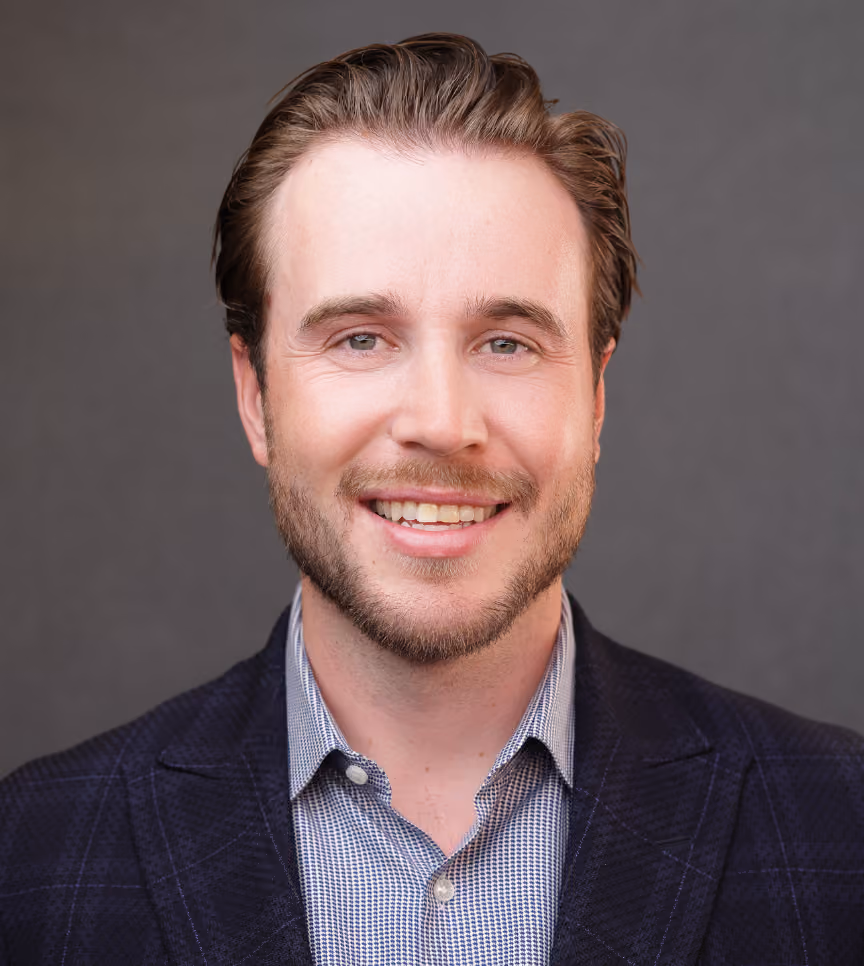 Smiling man with short brown hair, beard, wearing a checked shirt and dark blazer, gray background.