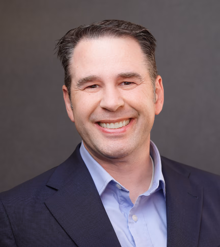 Smiling man with short dark hair wearing a navy blazer and light blue collared shirt on a gray background.