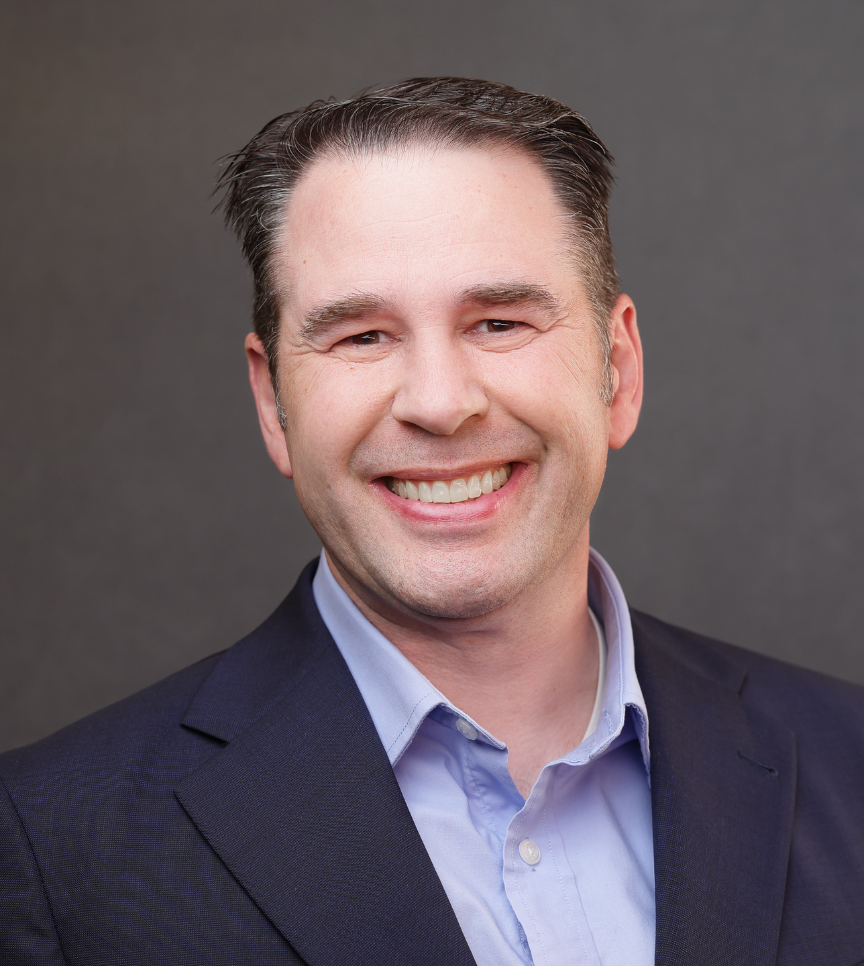 Smiling man with short dark hair wearing a navy blazer and light blue collared shirt on a gray background.