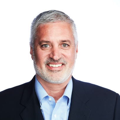 Smiling middle-aged man with gray hair, beard, wearing a blue shirt and dark blazer on white background.