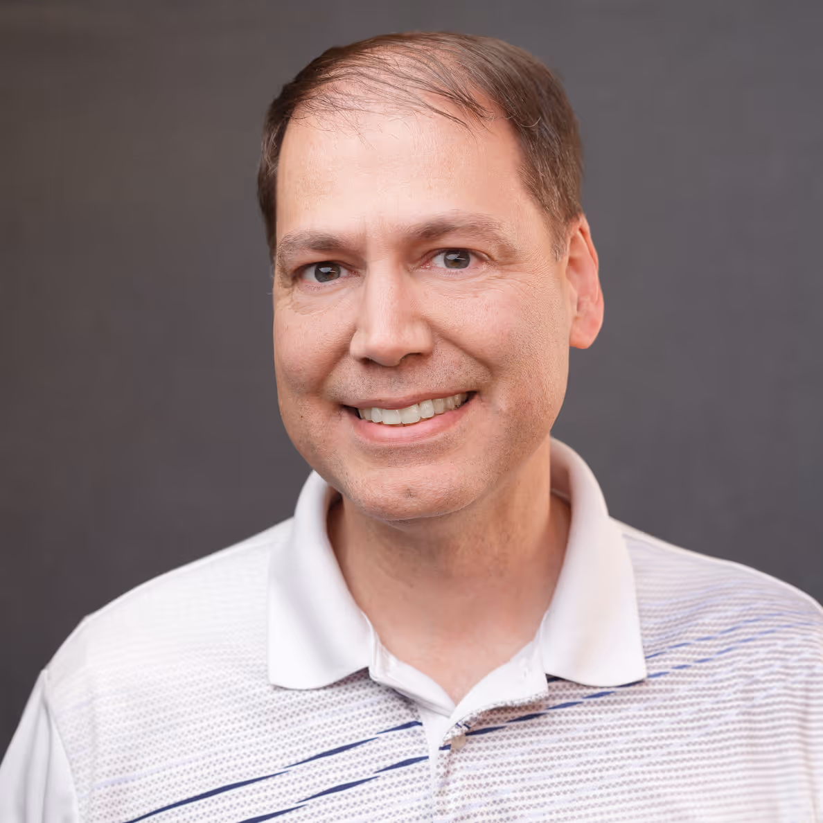 Smiling man with thinning brown hair wearing a white polo shirt against a gray background.