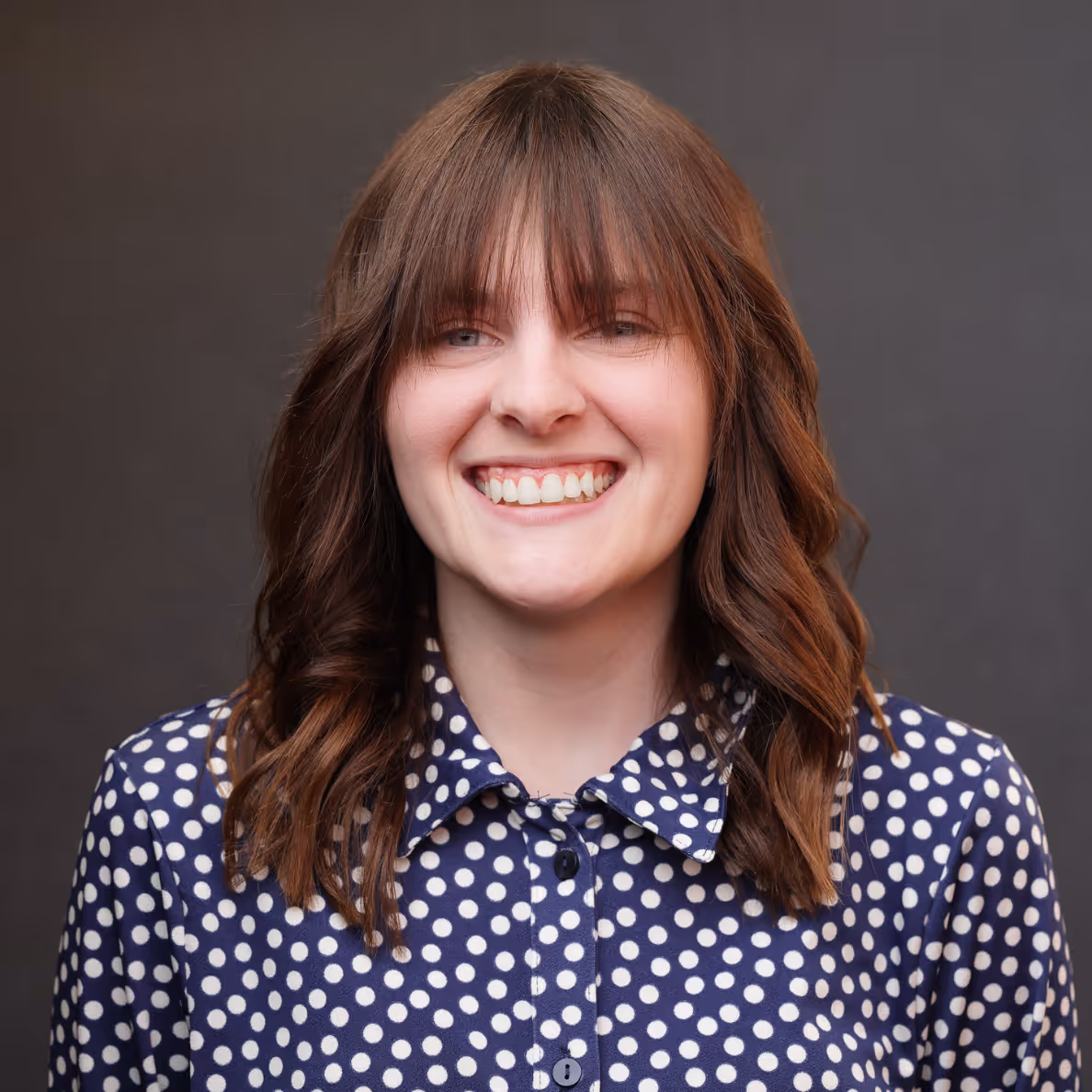 Smiling woman with shoulder-length brown hair and bangs wearing a navy blue polka dot shirt.