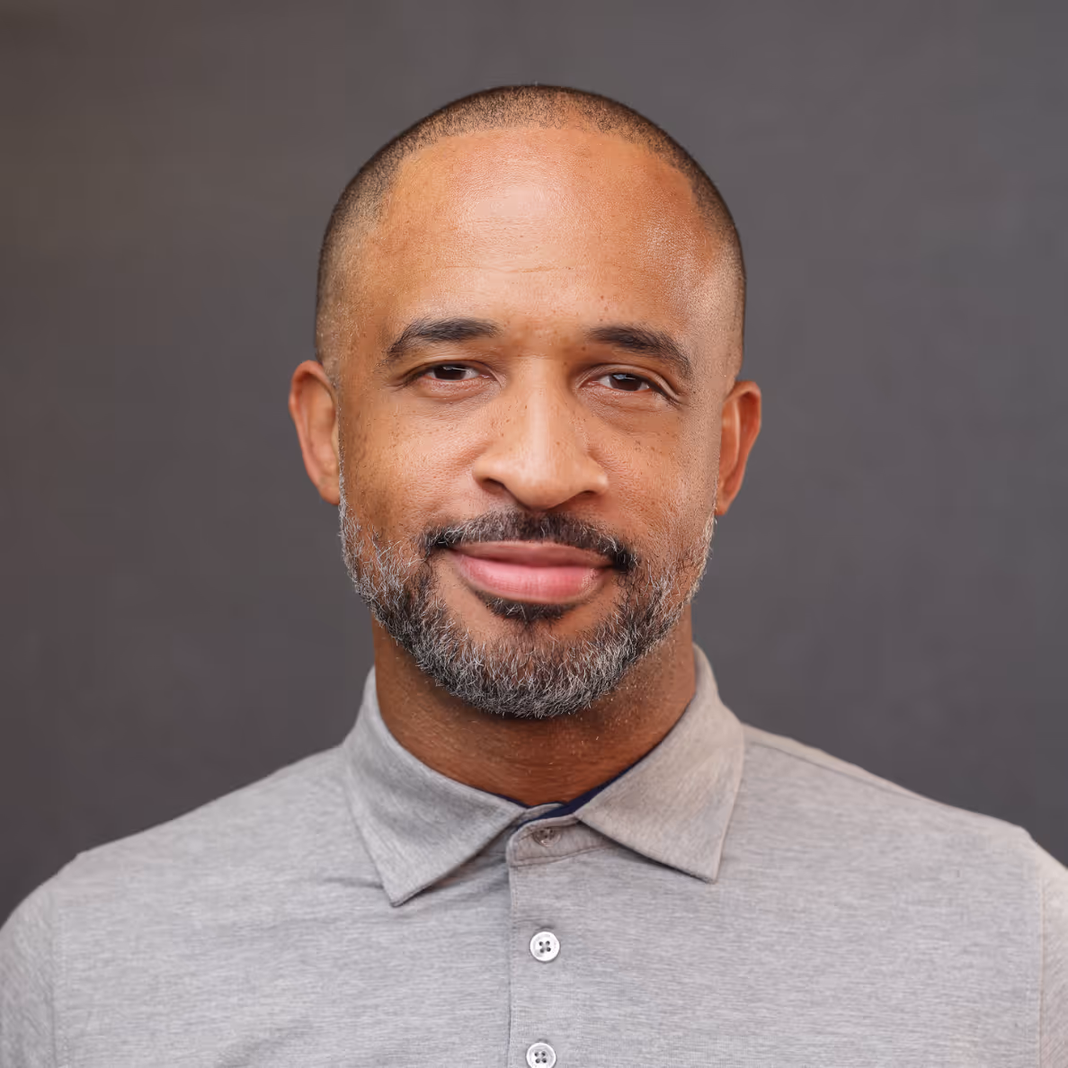 Smiling middle-aged man with short hair and gray beard wearing a light gray buttoned shirt.