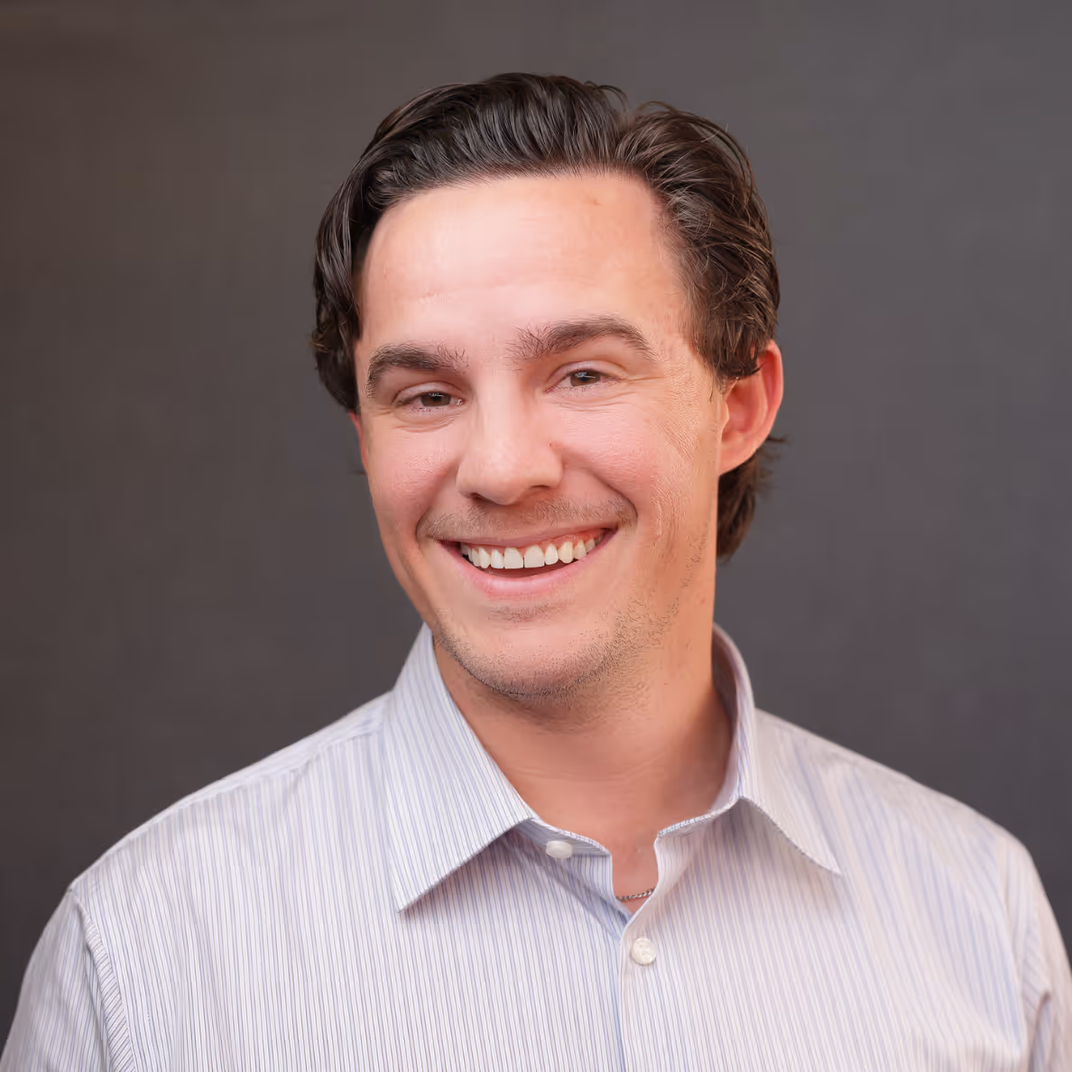 Smiling man with dark hair wearing a light striped shirt against a plain gray background.