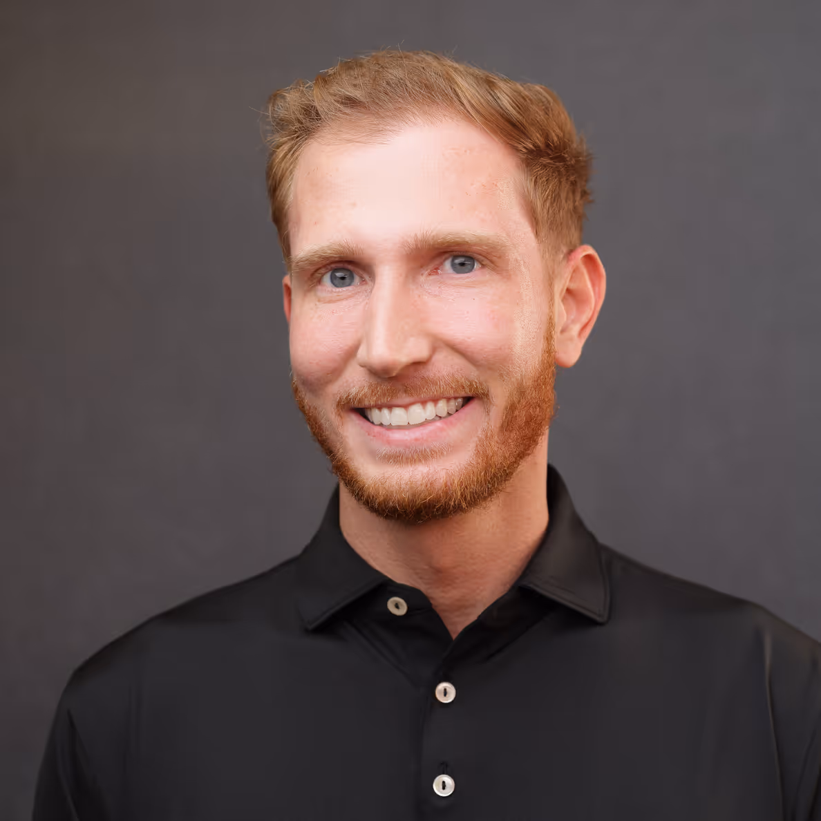 Smiling man with red hair and beard wearing a black buttoned shirt against a gray background.