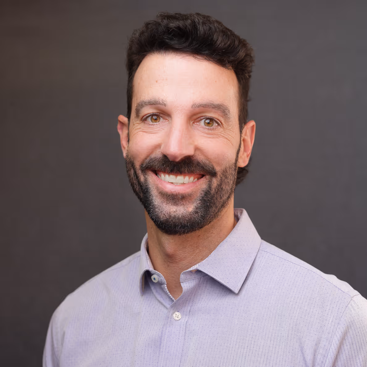 Smiling man with dark curly hair, beard, and light purple shirt against a gray background.