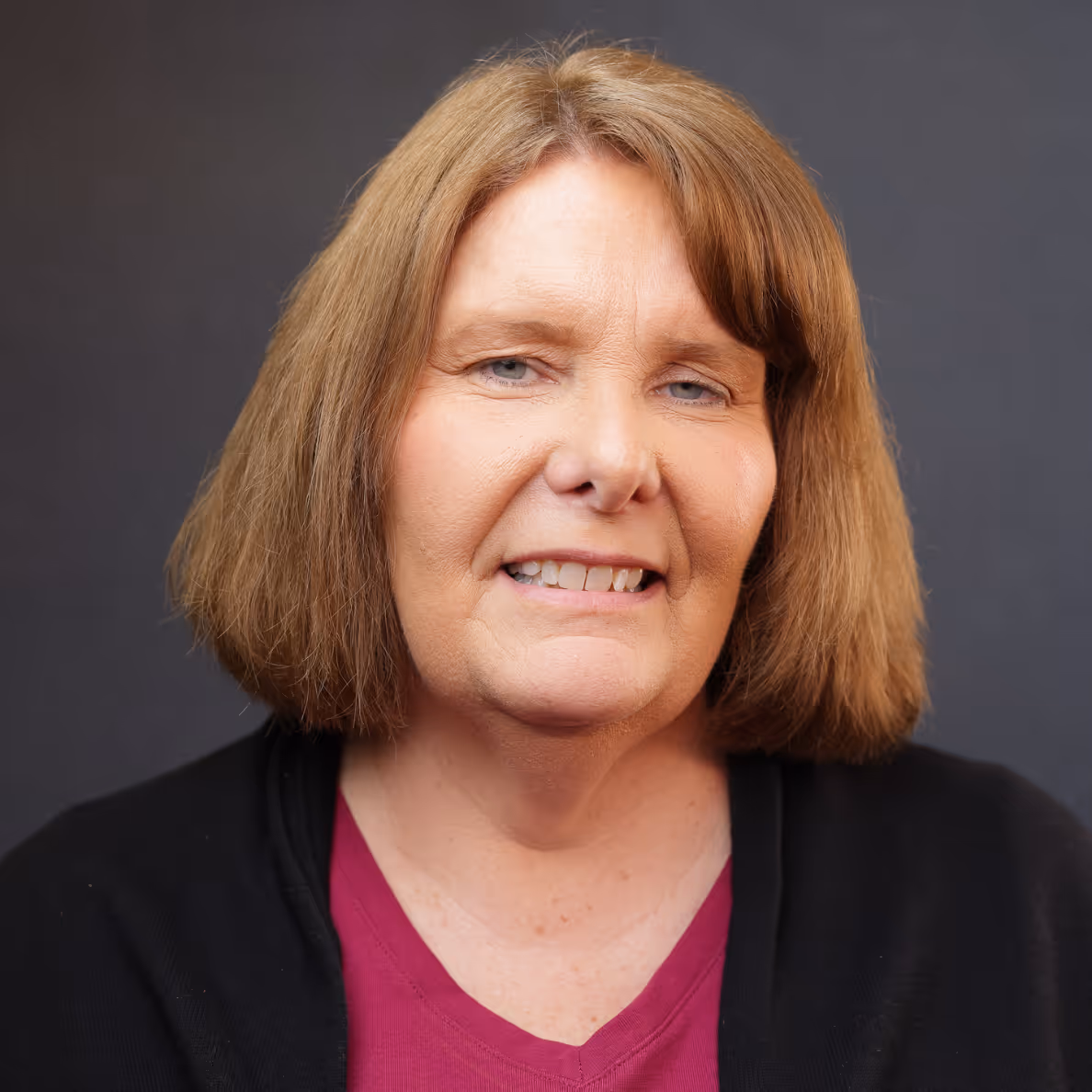Woman with light brown shoulder-length hair wearing a maroon top and black jacket smiling slightly.