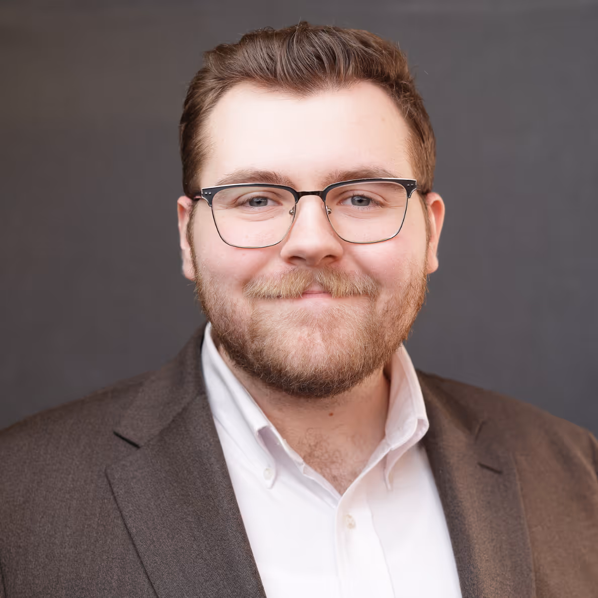 Smiling man with glasses, beard, and mustache wearing a brown suit jacket and white shirt.