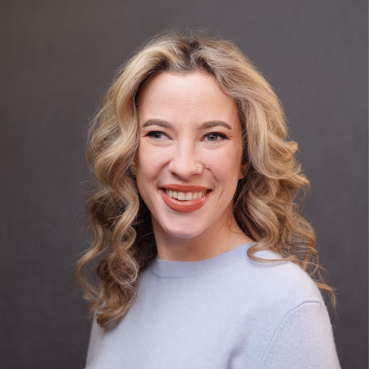 Smiling woman with curly blond hair wearing a light blue sweater against a gray background.