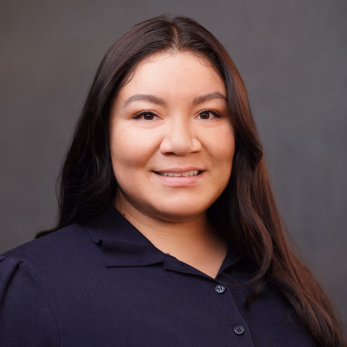 Smiling woman with long dark hair wearing a navy blue collared shirt against a gray background.