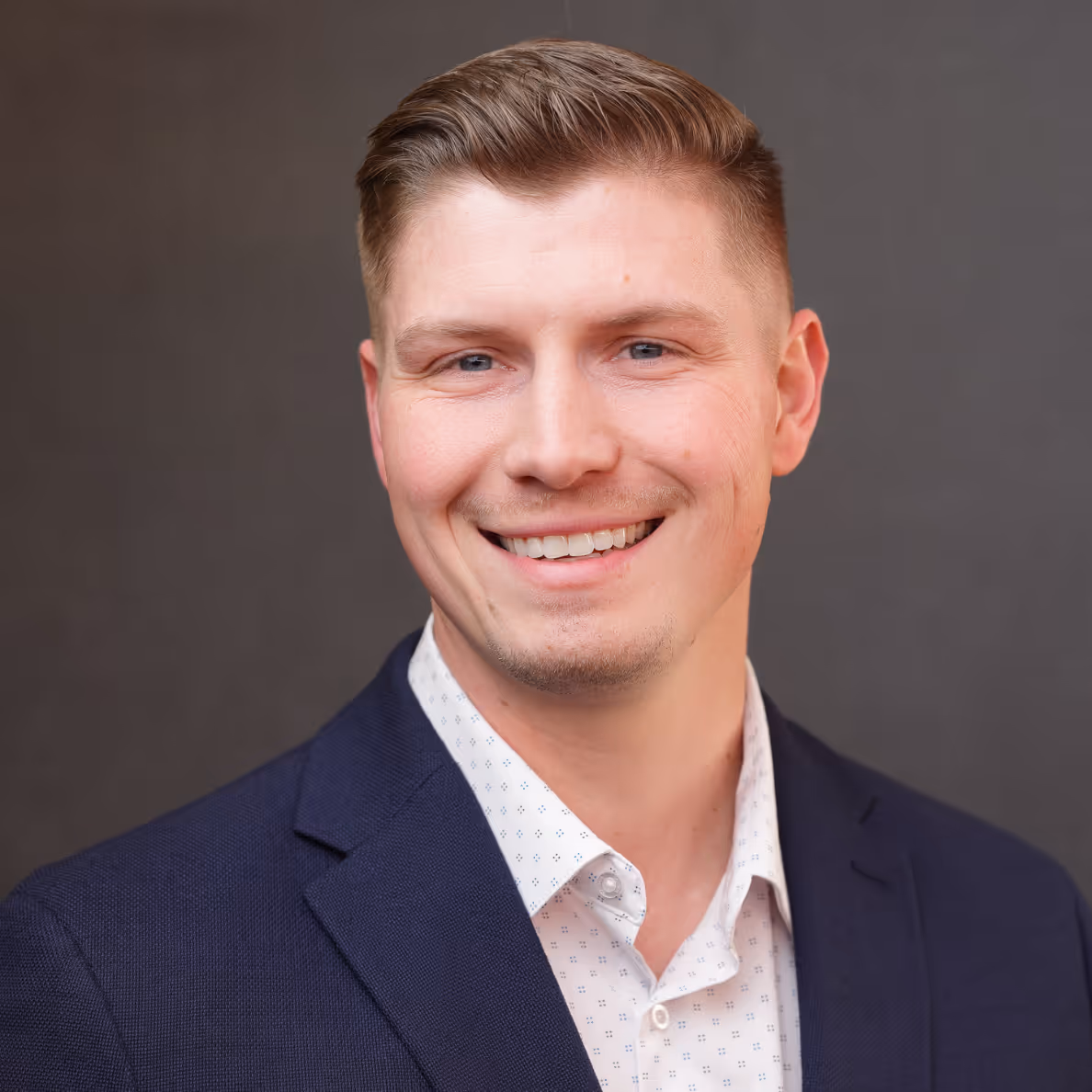 Smiling man with short light brown hair wearing a navy blazer and white patterned shirt.
