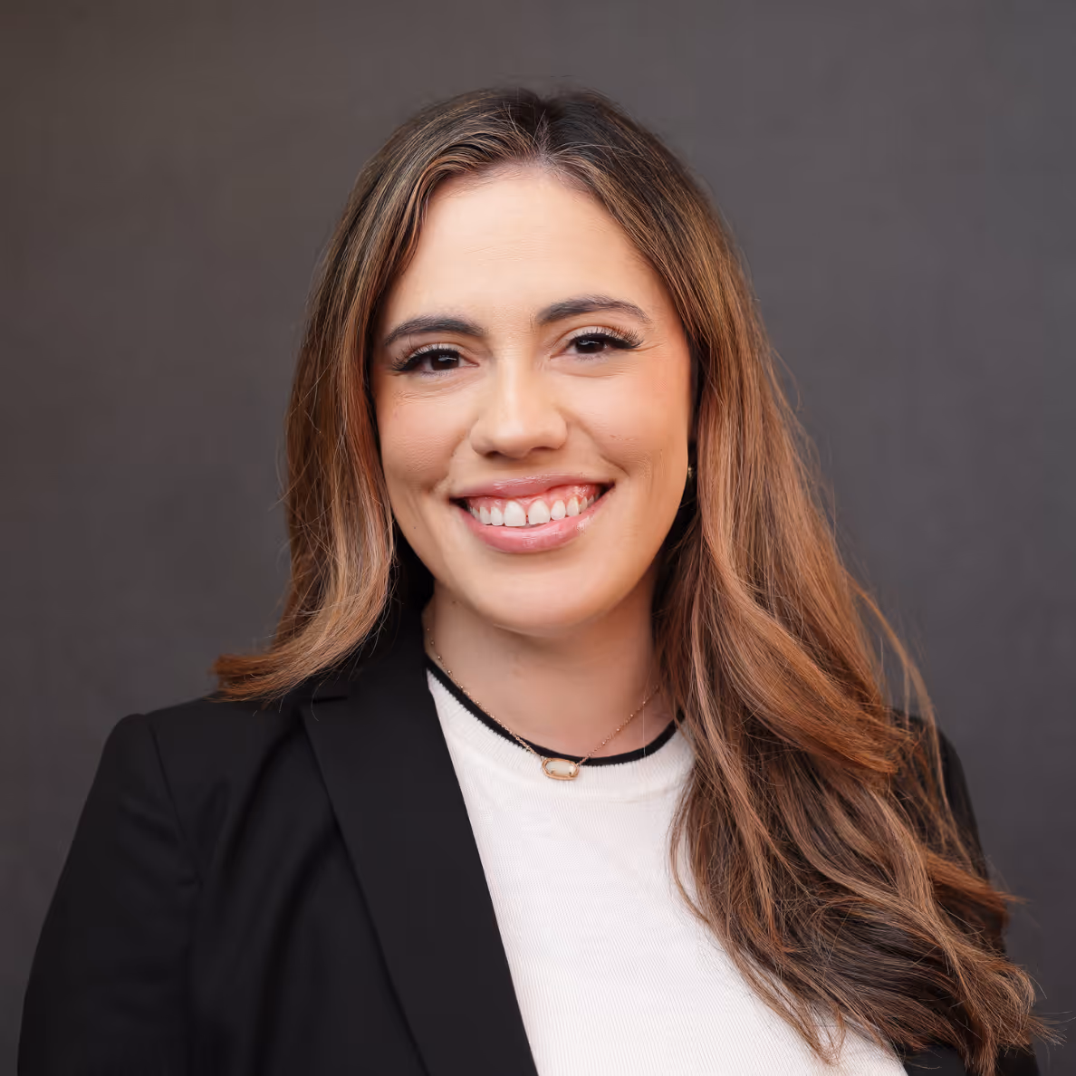 Smiling woman with long brown hair wearing a black blazer and white top against a gray background.