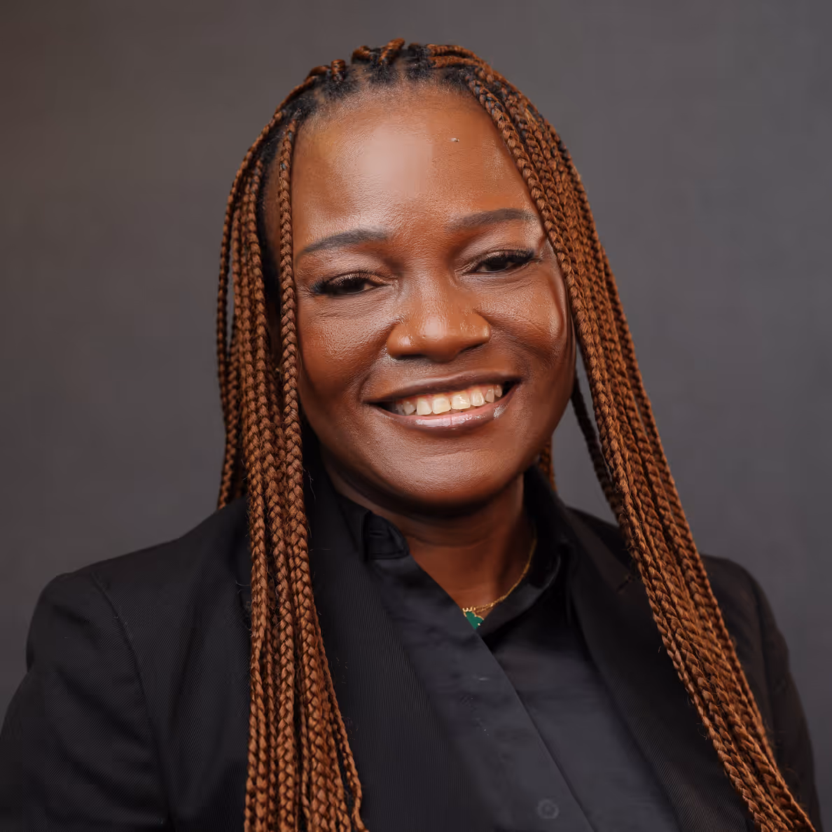 Smiling woman with long braided hair wearing a black jacket and shirt against a gray background.