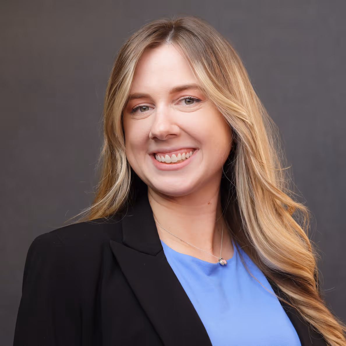 Smiling woman with long blonde hair wearing a black blazer and blue top against a gray background.