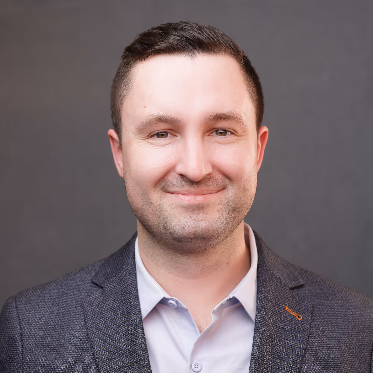 Smiling man with short dark hair in gray suit jacket and light shirt against gray background.