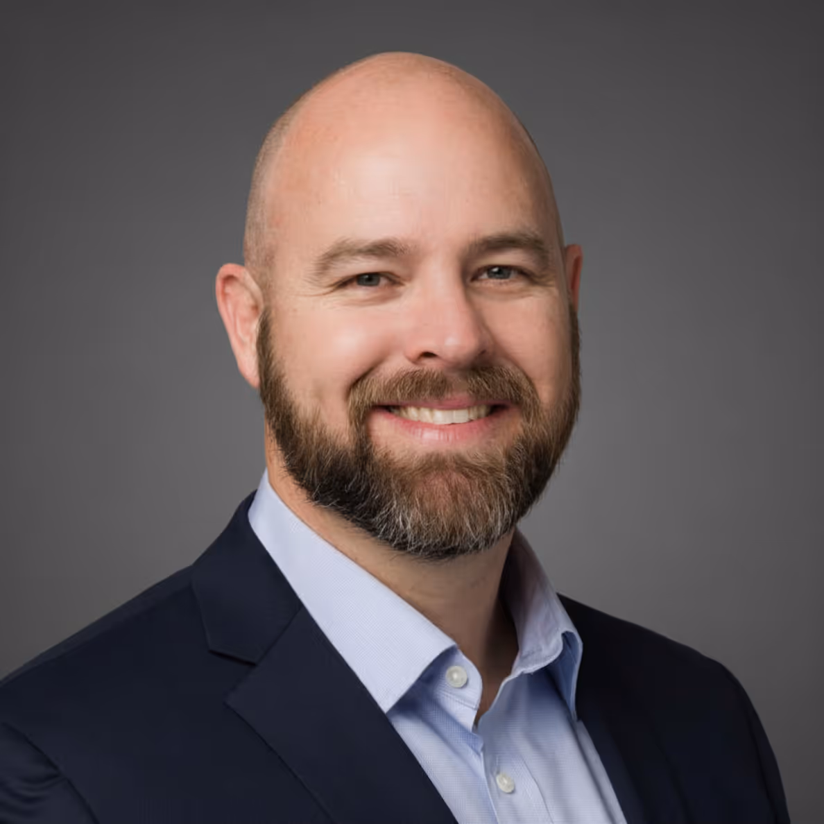 Smiling bald man with a beard wearing a blue suit jacket and light blue shirt on gray background.