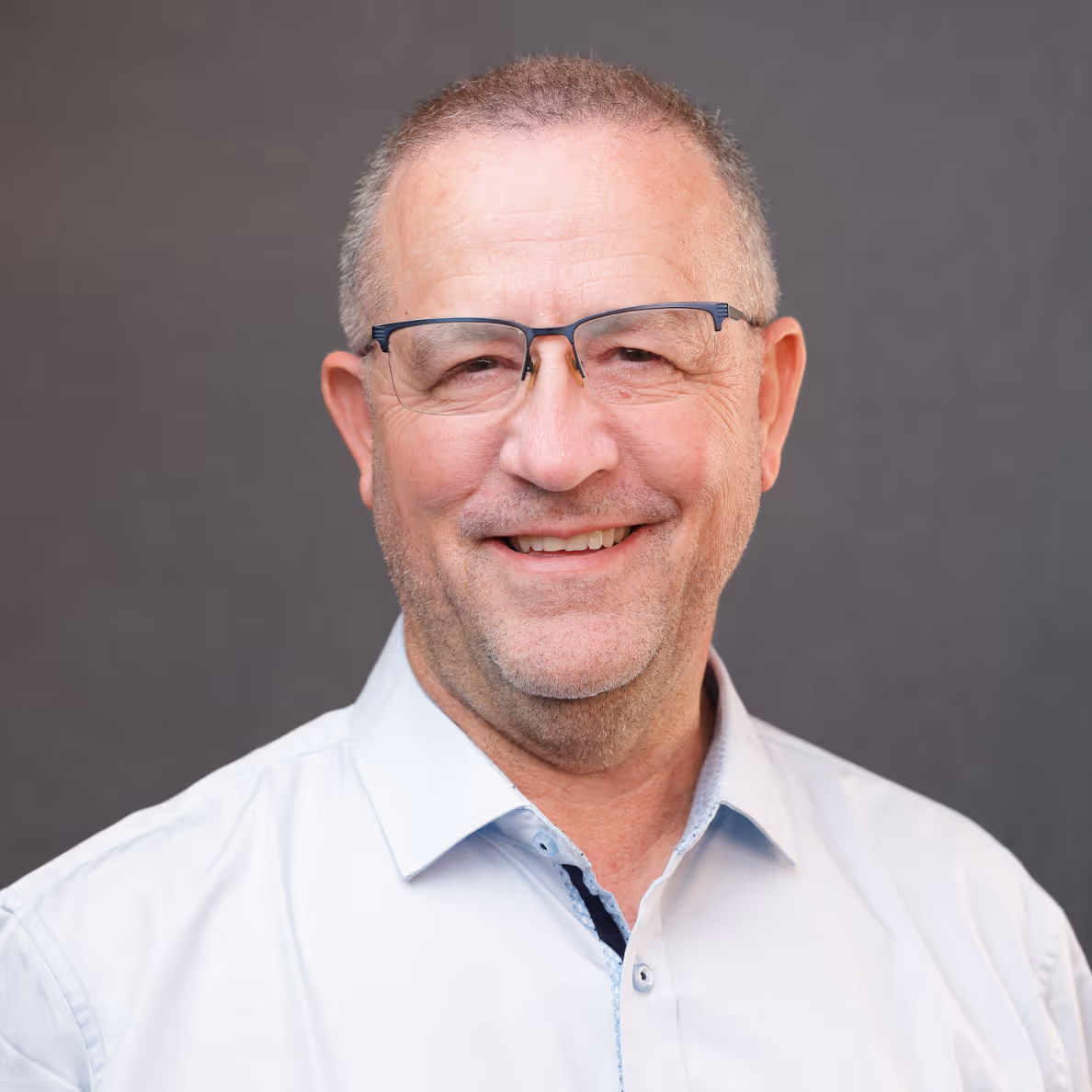 Smiling middle-aged man with glasses wearing a white button-up shirt on a gray background.