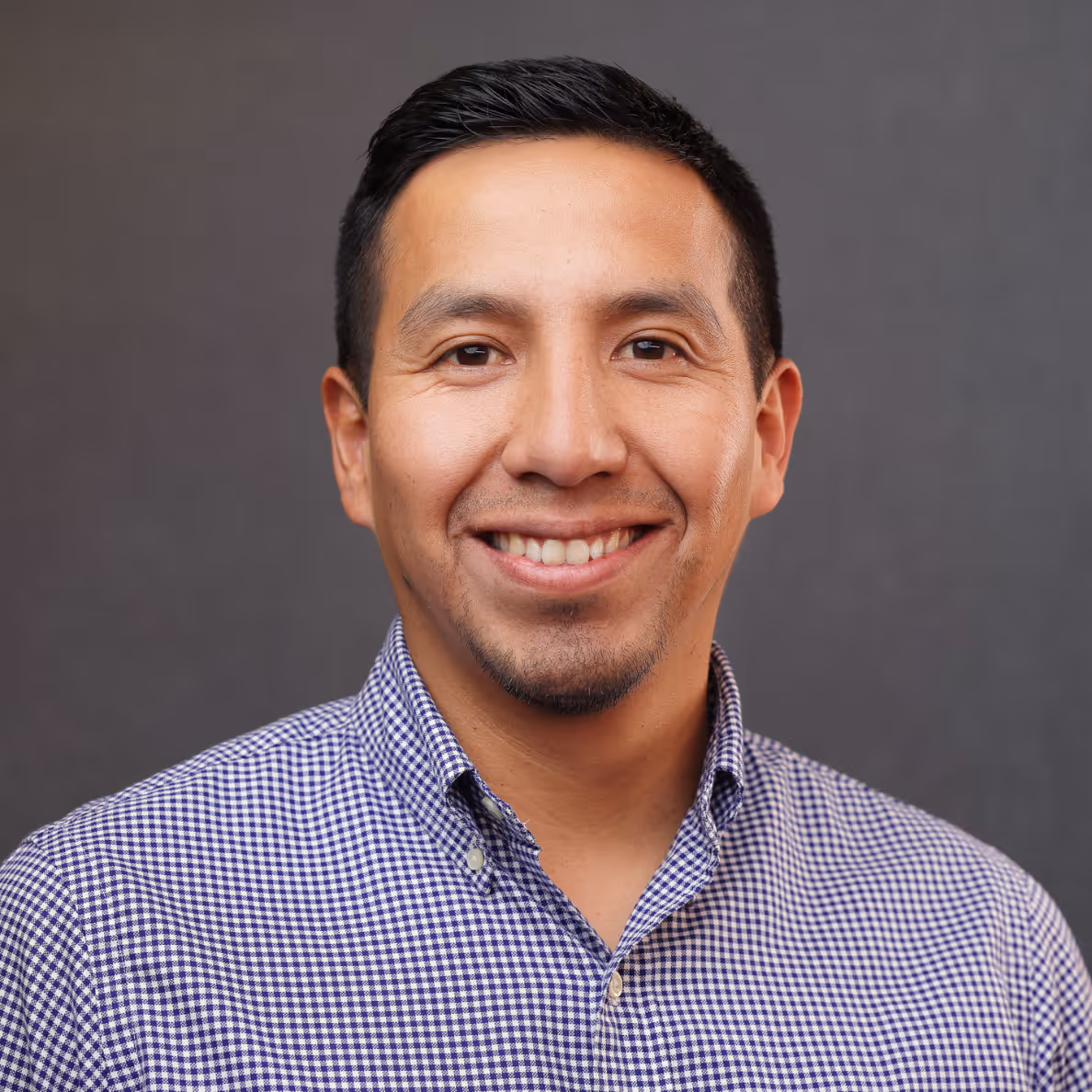 Smiling man with short black hair wearing a blue checkered button-up shirt against a gray background.
