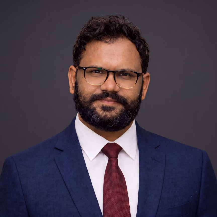 Man with curly hair, beard, glasses, navy suit, white shirt, and maroon tie against dark background.