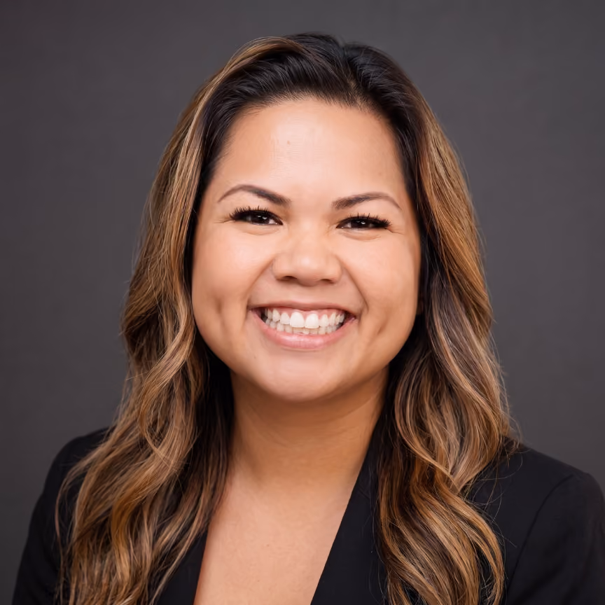 Smiling woman with long wavy hair wearing a black blazer against a gray background.