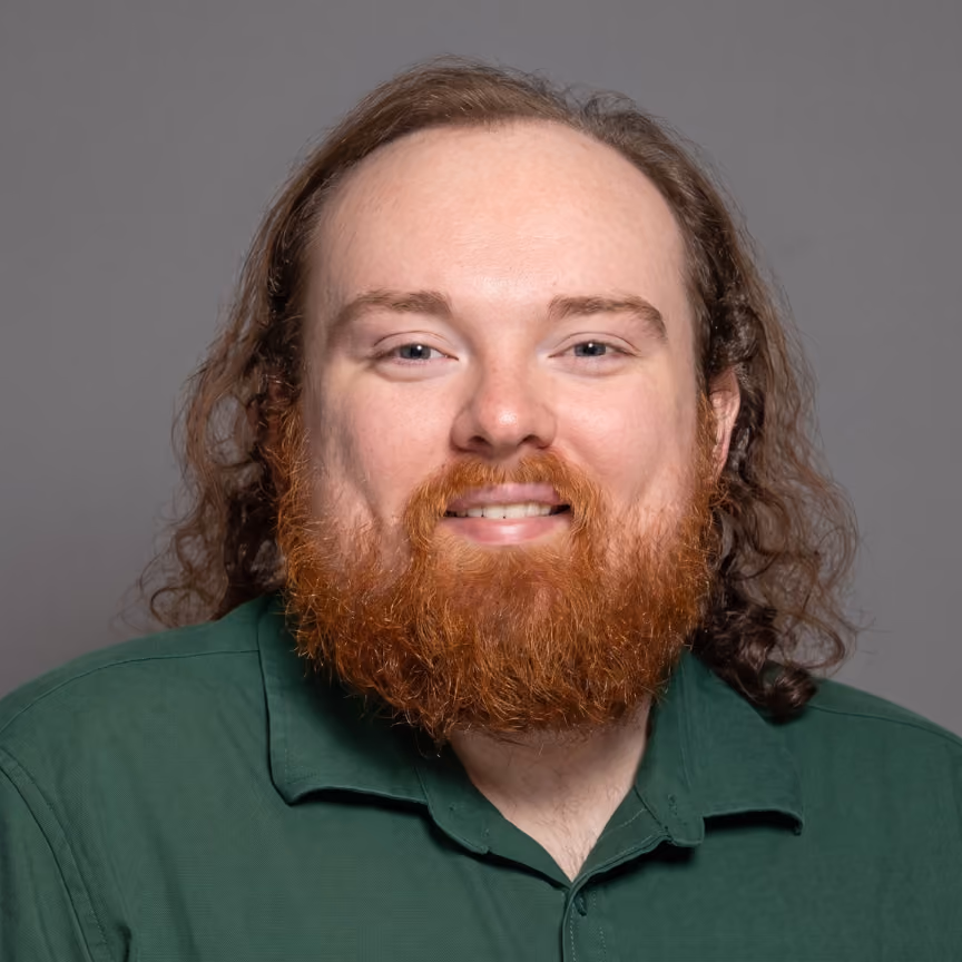 Smiling man with long curly hair and reddish beard wearing a green collared shirt on gray background.