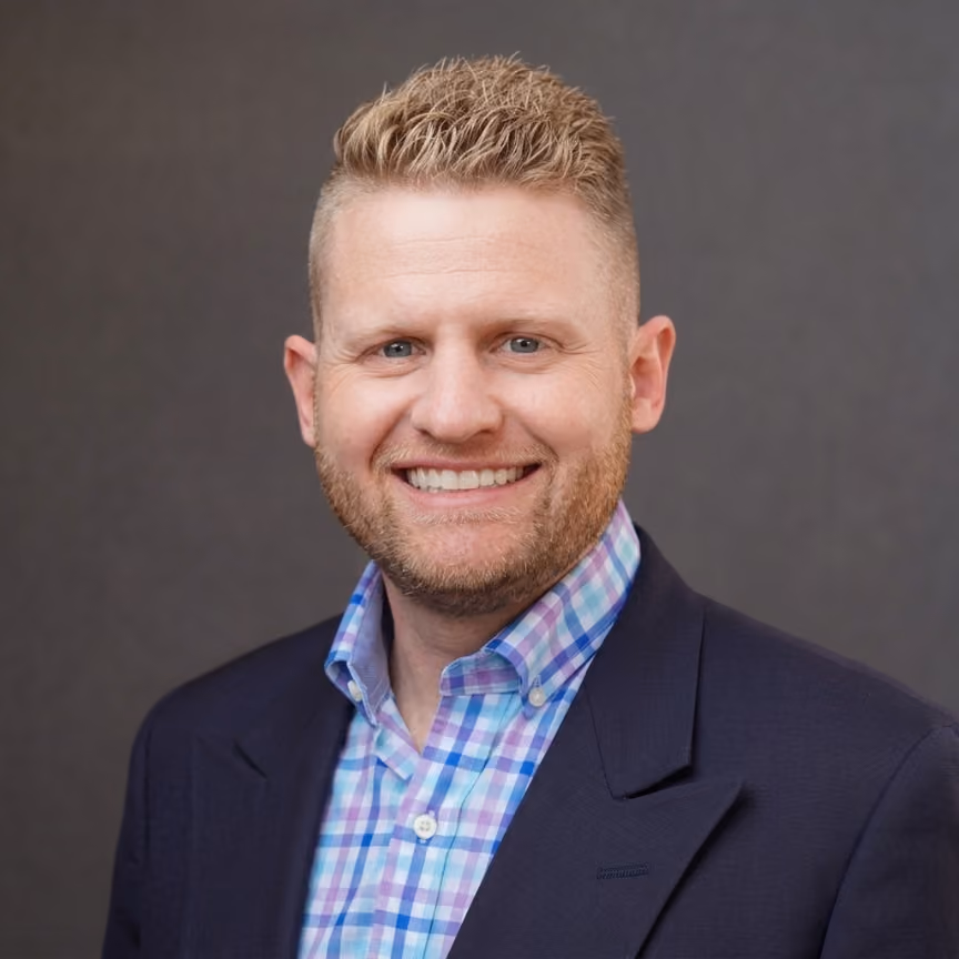 Smiling man with short blond hair, beard, wearing a plaid shirt and dark blazer against gray background.