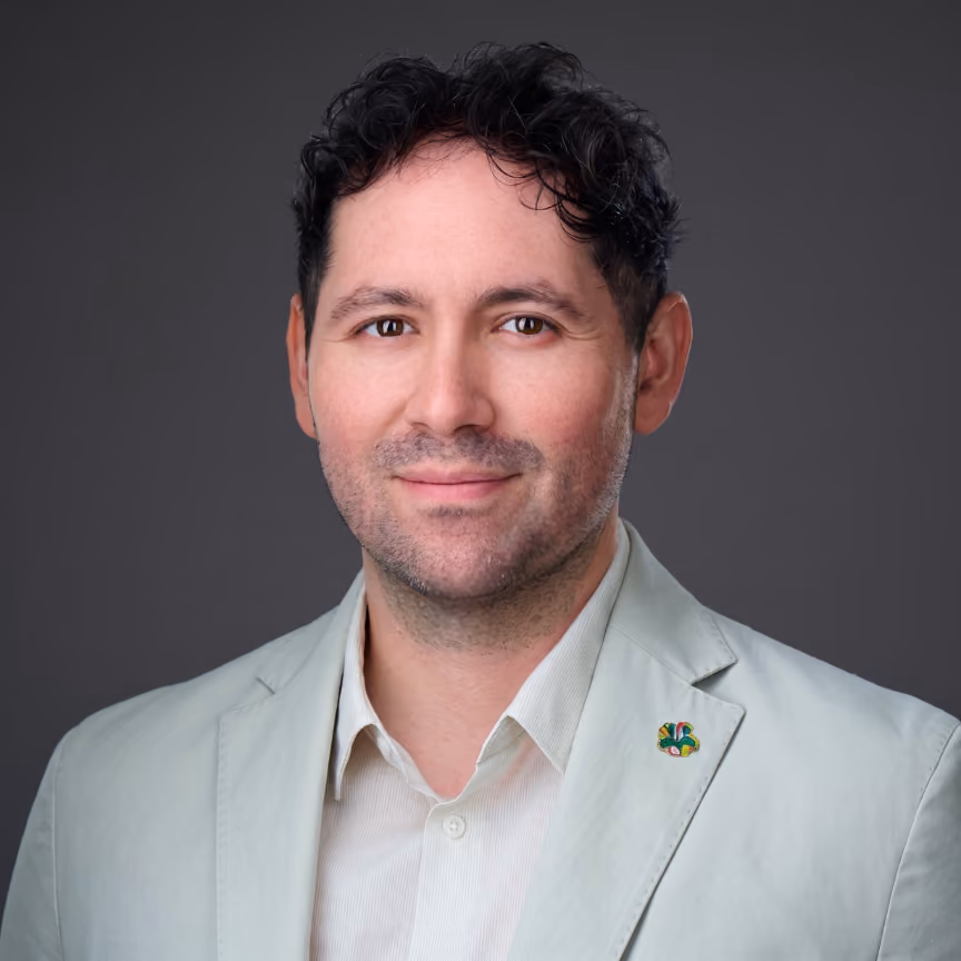Man with short curly black hair wearing a light gray blazer and white shirt smiling slightly, gray background.