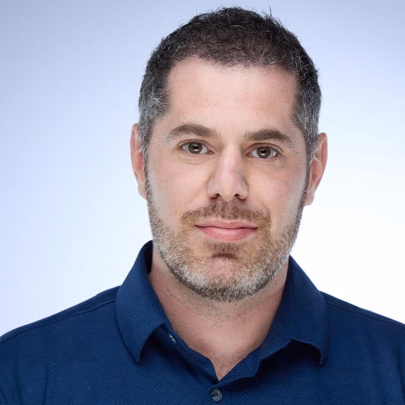Close-up of a man with short dark hair and a trimmed beard wearing a blue collared shirt.