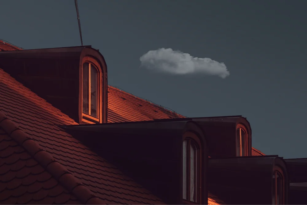 Rooftop with red tiles and dormer windows illuminated by warm light under a dark sky with a single white cloud.