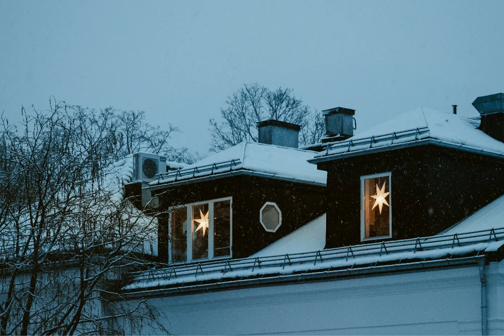 Snow-covered rooftops of dark houses at dusk with illuminated star decorations visible through two windows.
