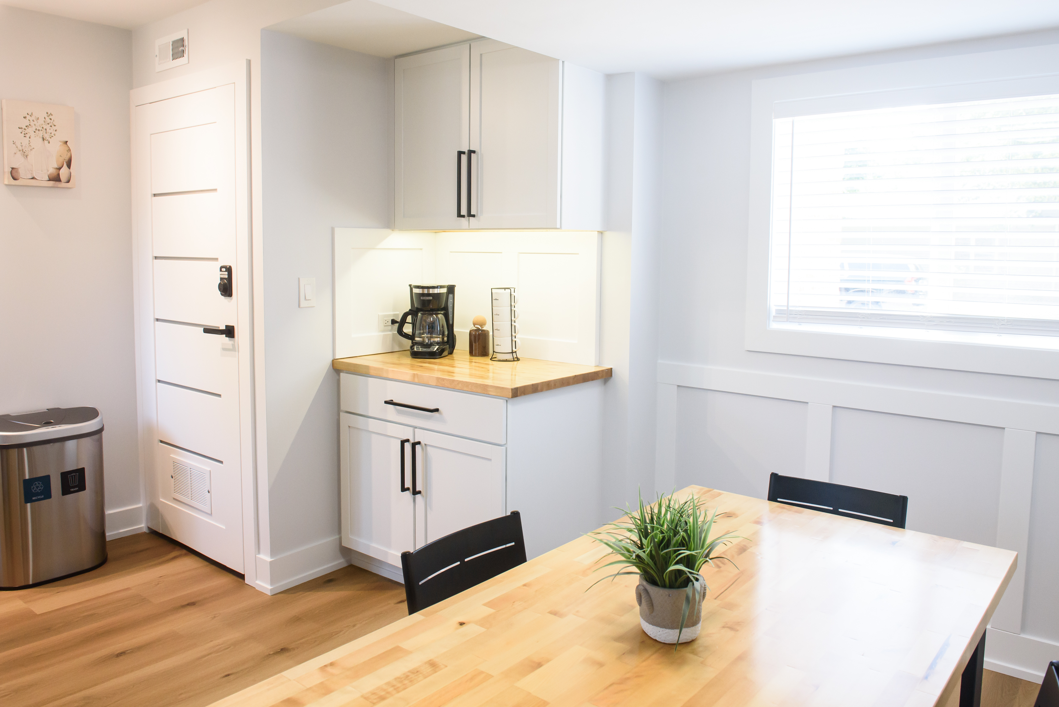 Bright kitchen corner with white cabinets, coffee maker, spice rack, wooden table with potted plant, and window with blinds.