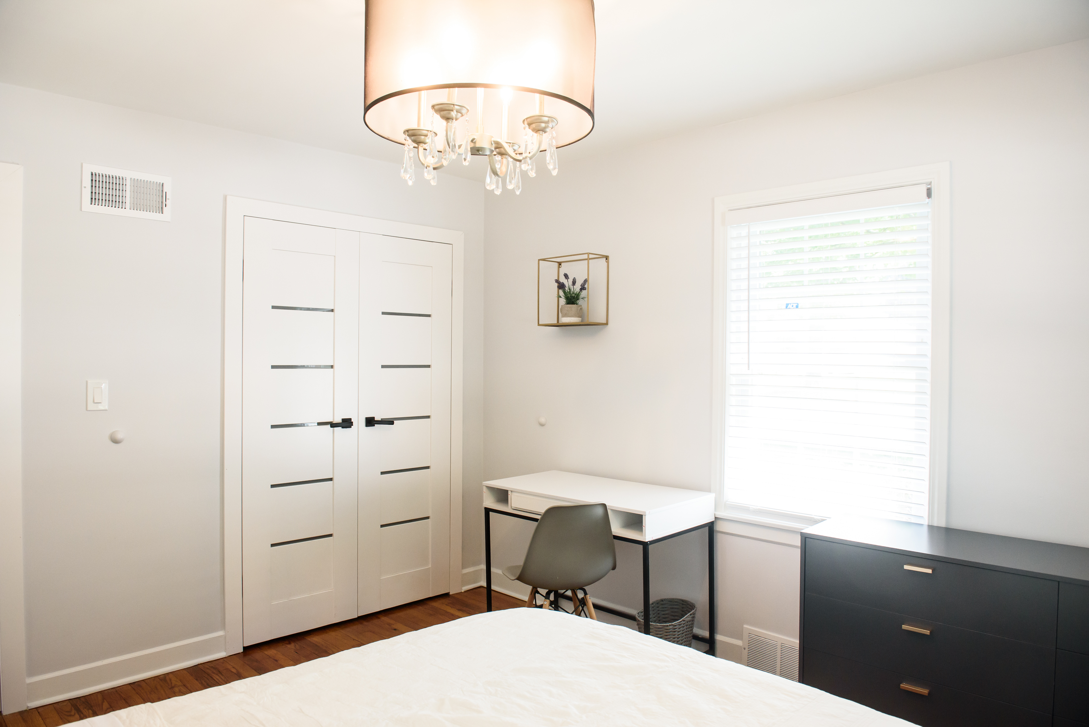 Bright bedroom corner with white double doors, a white desk with a gray chair, a black dresser, and a window with blinds.