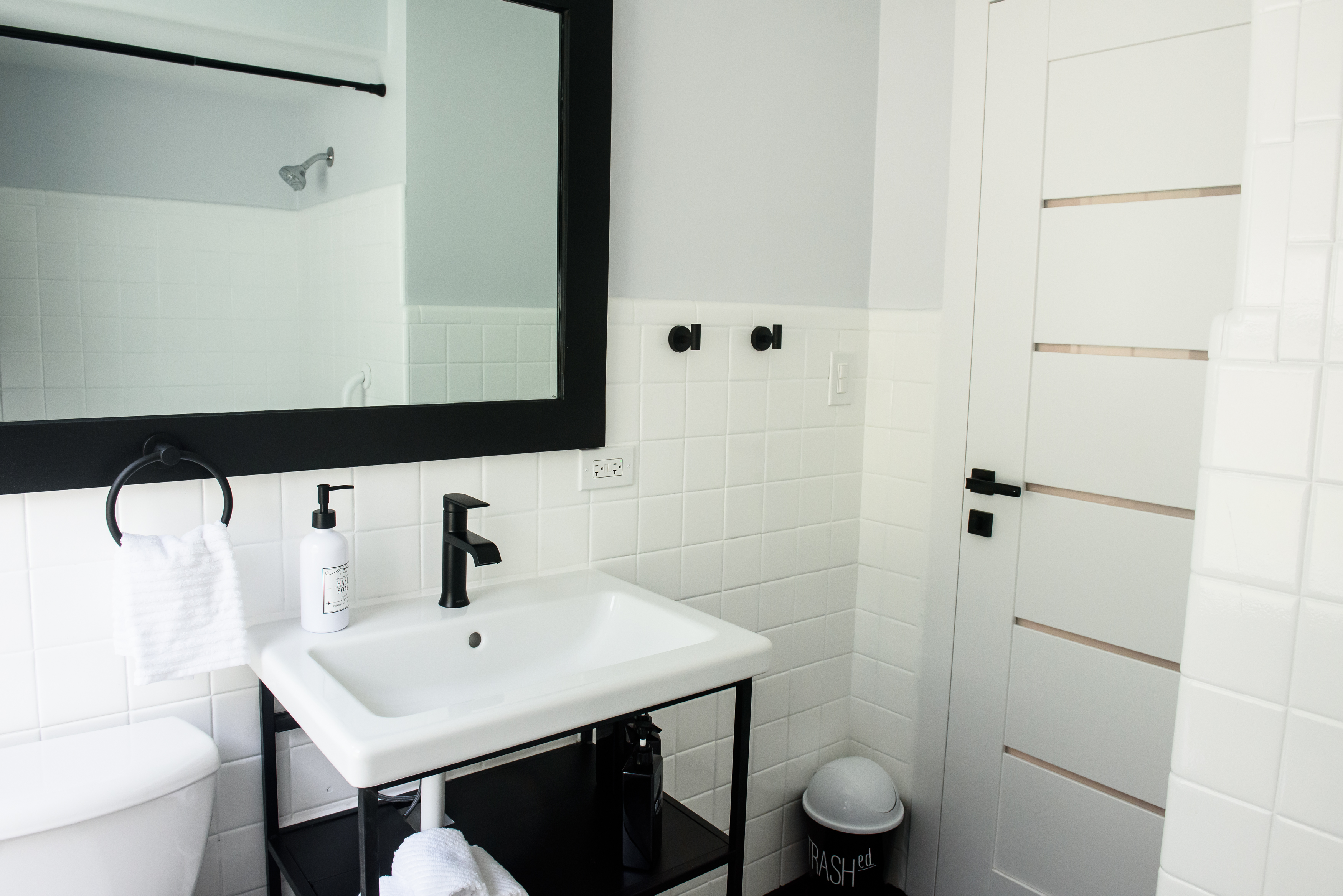 Minimalist bathroom sink with black faucet, a large black-framed mirror, white tiled walls, a towel ring with a white towel, soap dispenser, and a trash bin.