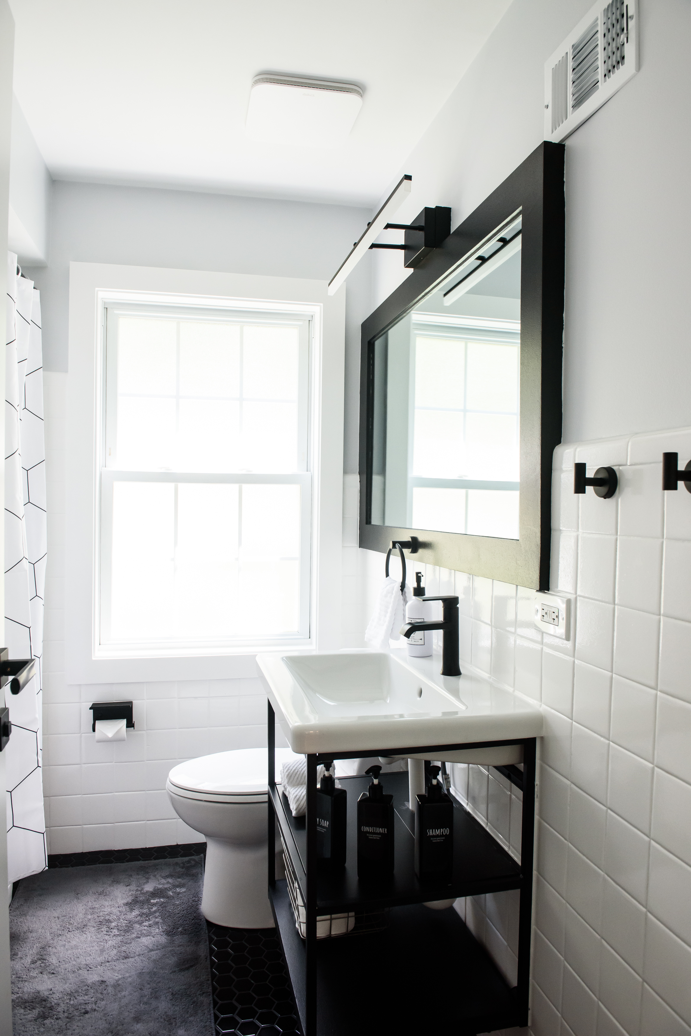 Bright modern bathroom with white tiles, black-framed mirror, white sink on a black metal stand, and a white toilet under a window.