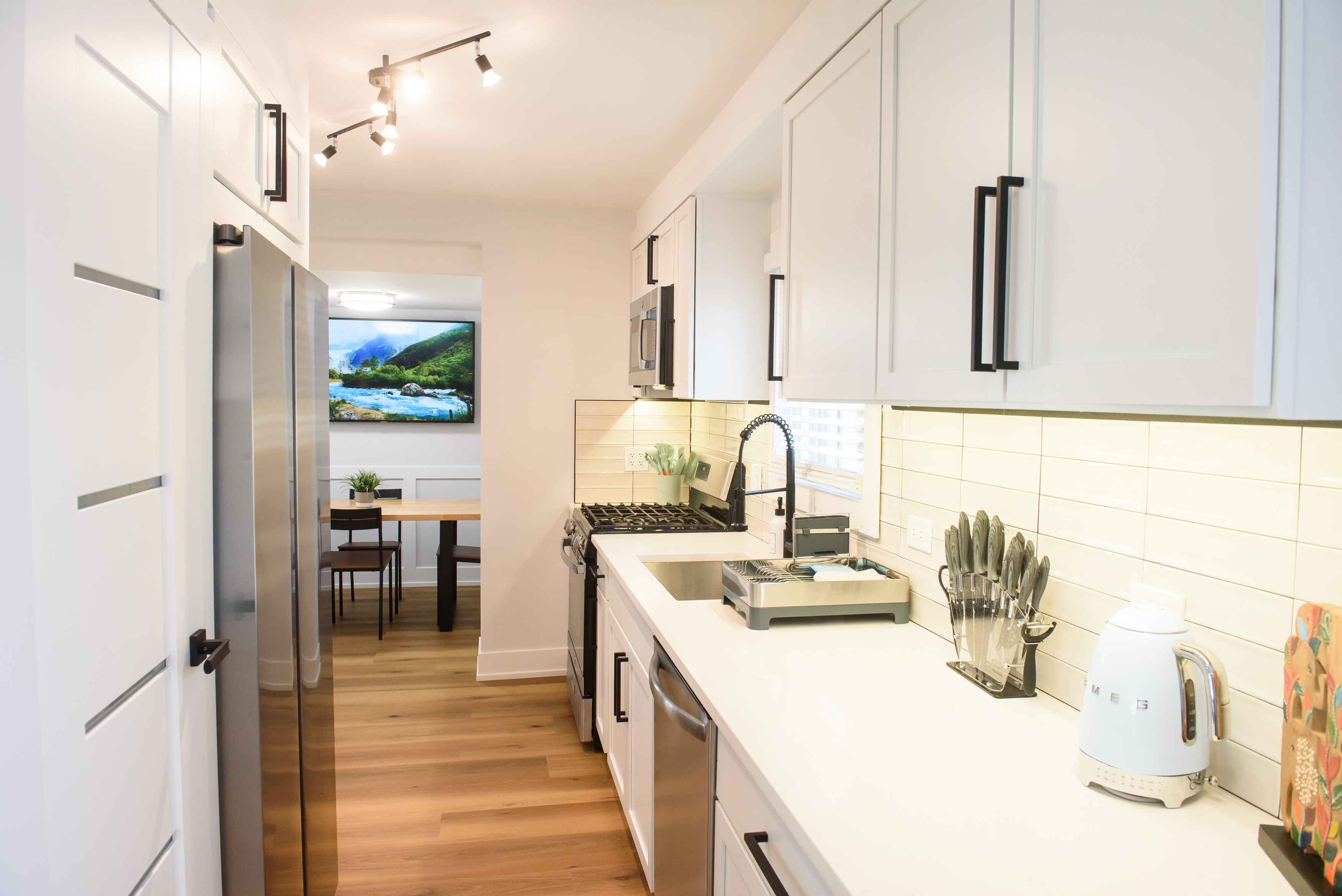 Modern kitchen with white cabinets, stainless steel appliances, a sink, knife block, electric kettle, and a view into a dining area with a mounted TV.