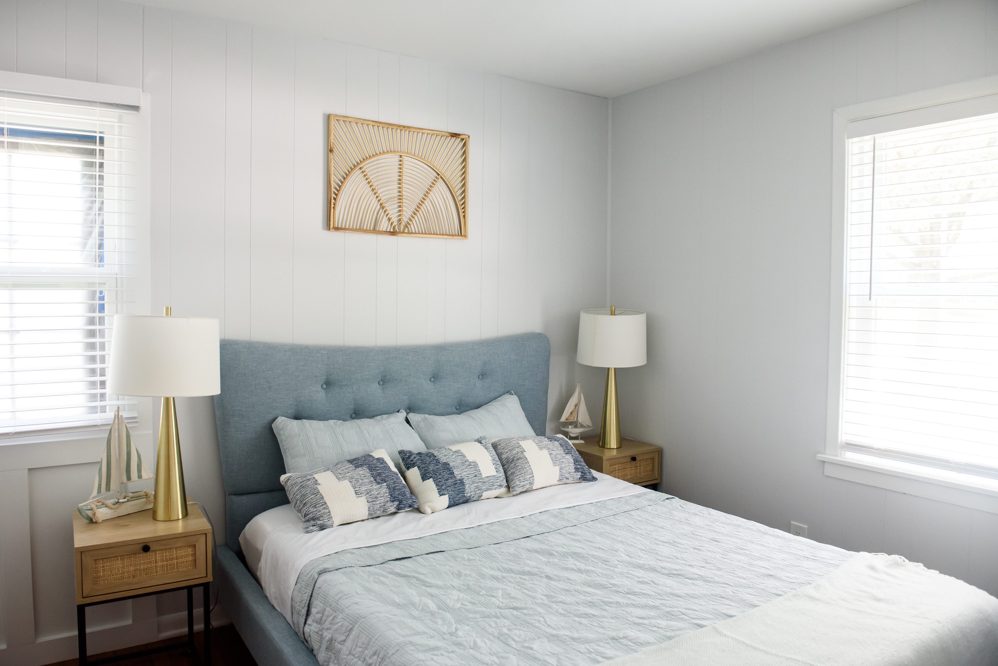 Bright bedroom with a blue upholstered bed, blue and white pillows, wooden nightstands with gold lamps, and a decorative wall hanging.