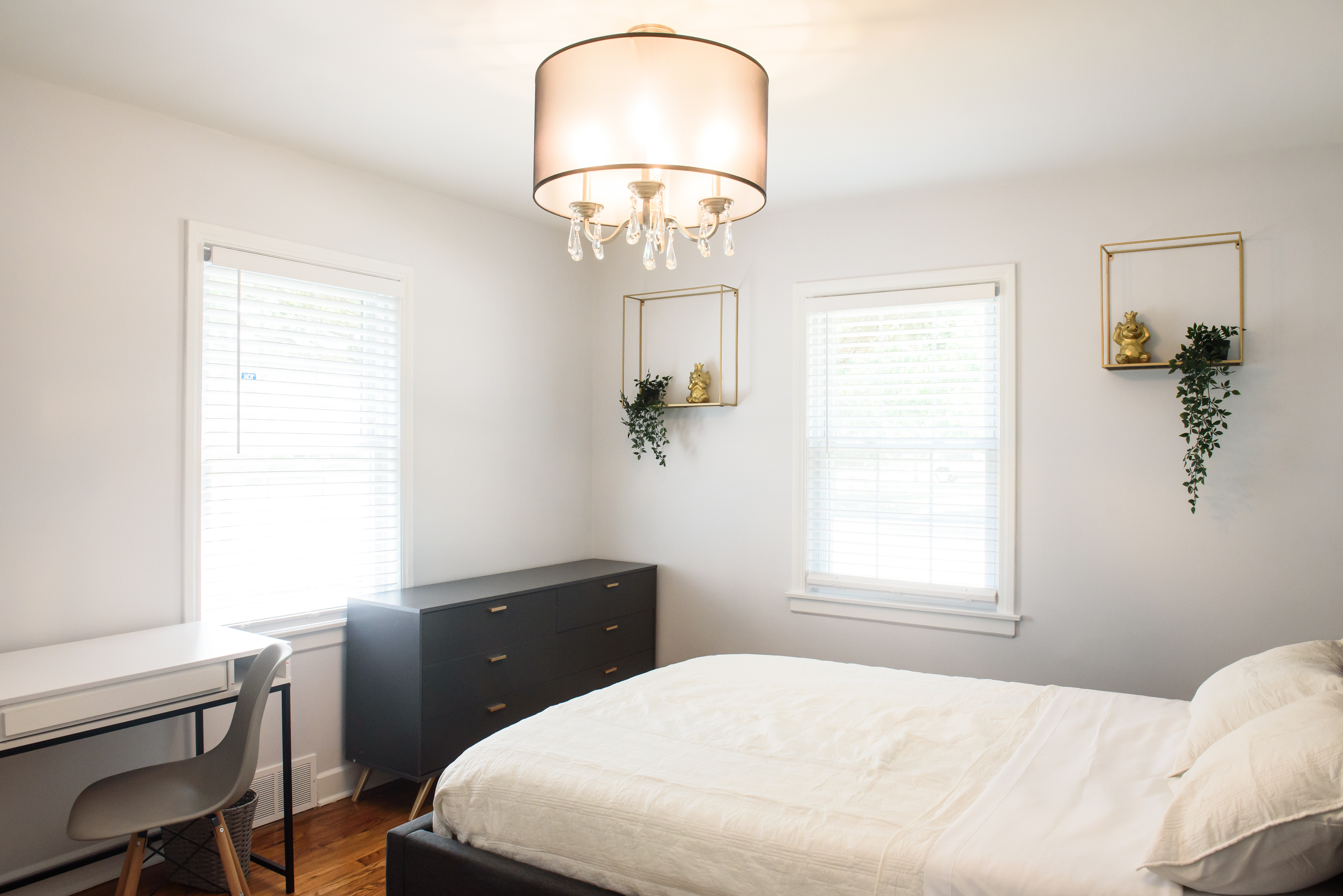Bright bedroom with white bedding, dark dresser, desk with chair, two windows with blinds, and hanging plants in gold-framed shelves.