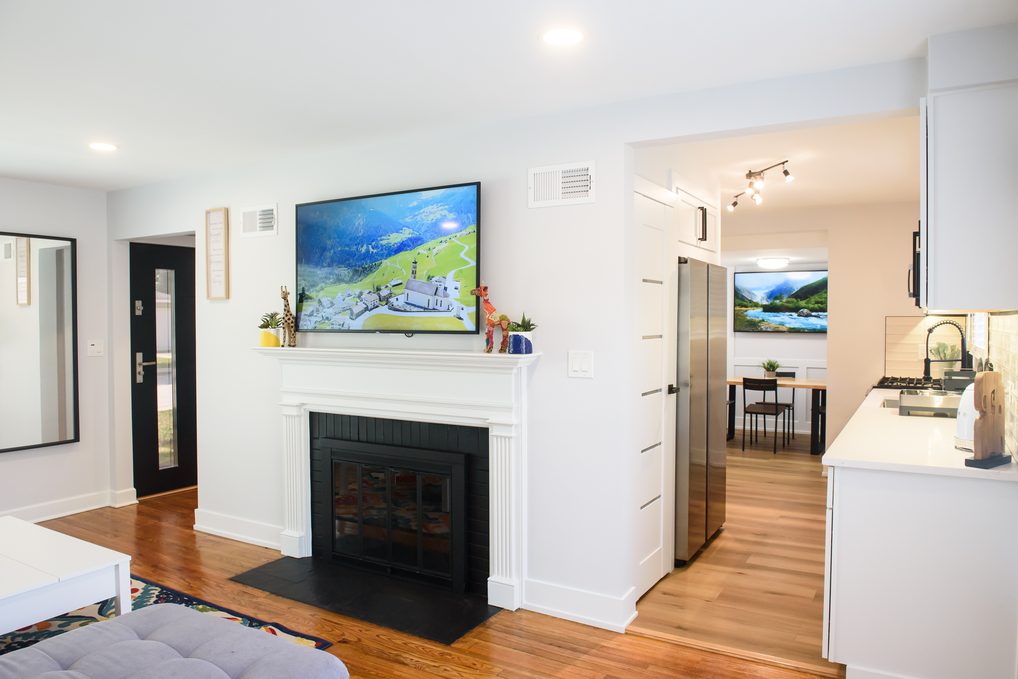 Bright living room with white walls, a white fireplace with TV above, hardwood floors, and a view into a kitchen with stainless steel appliances.