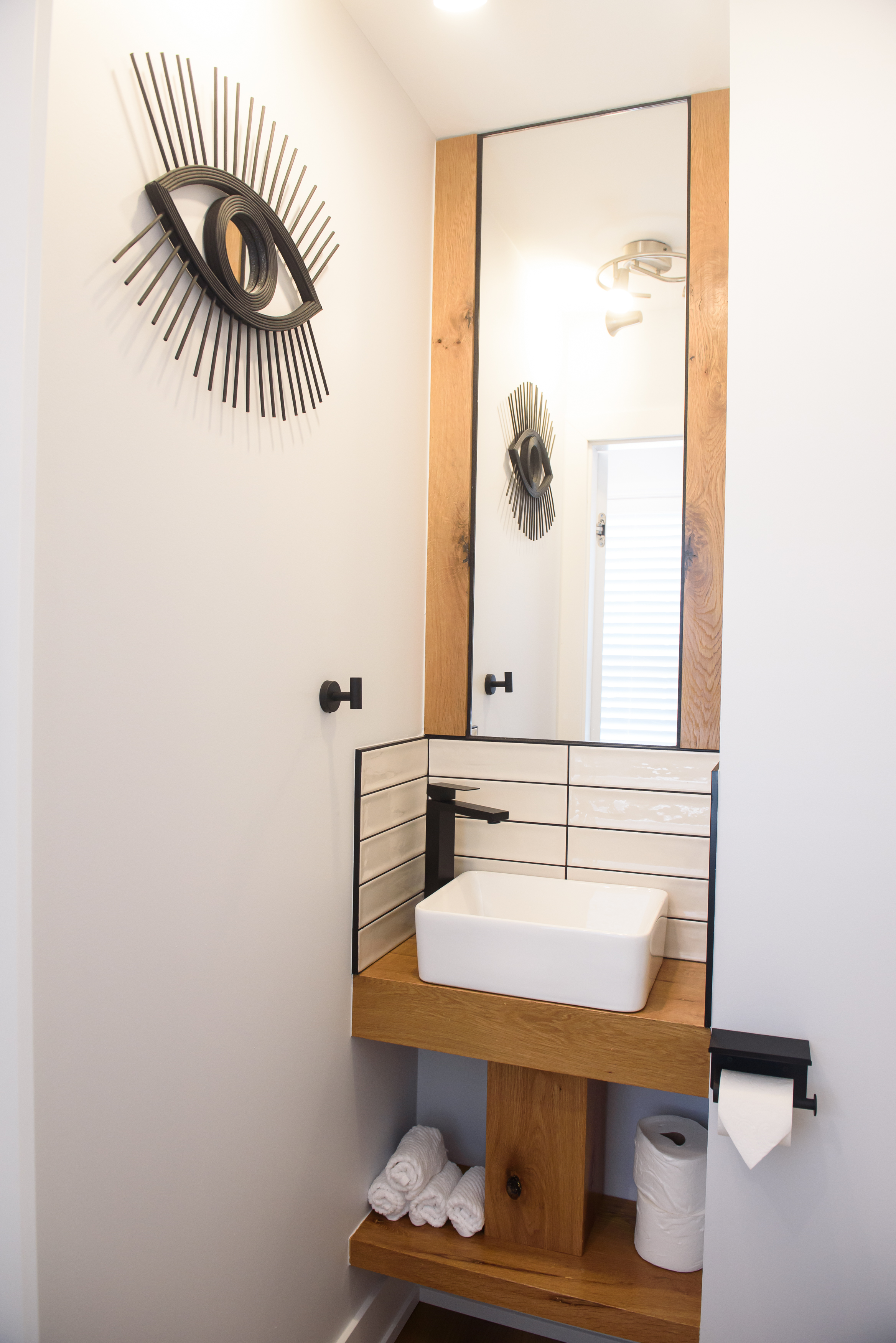 Small modern bathroom sink area with rectangular white vessel sink, black faucet, large mirror framed in wood, white tiled backsplash, and decorative black metal eye sculpture on the wall.
