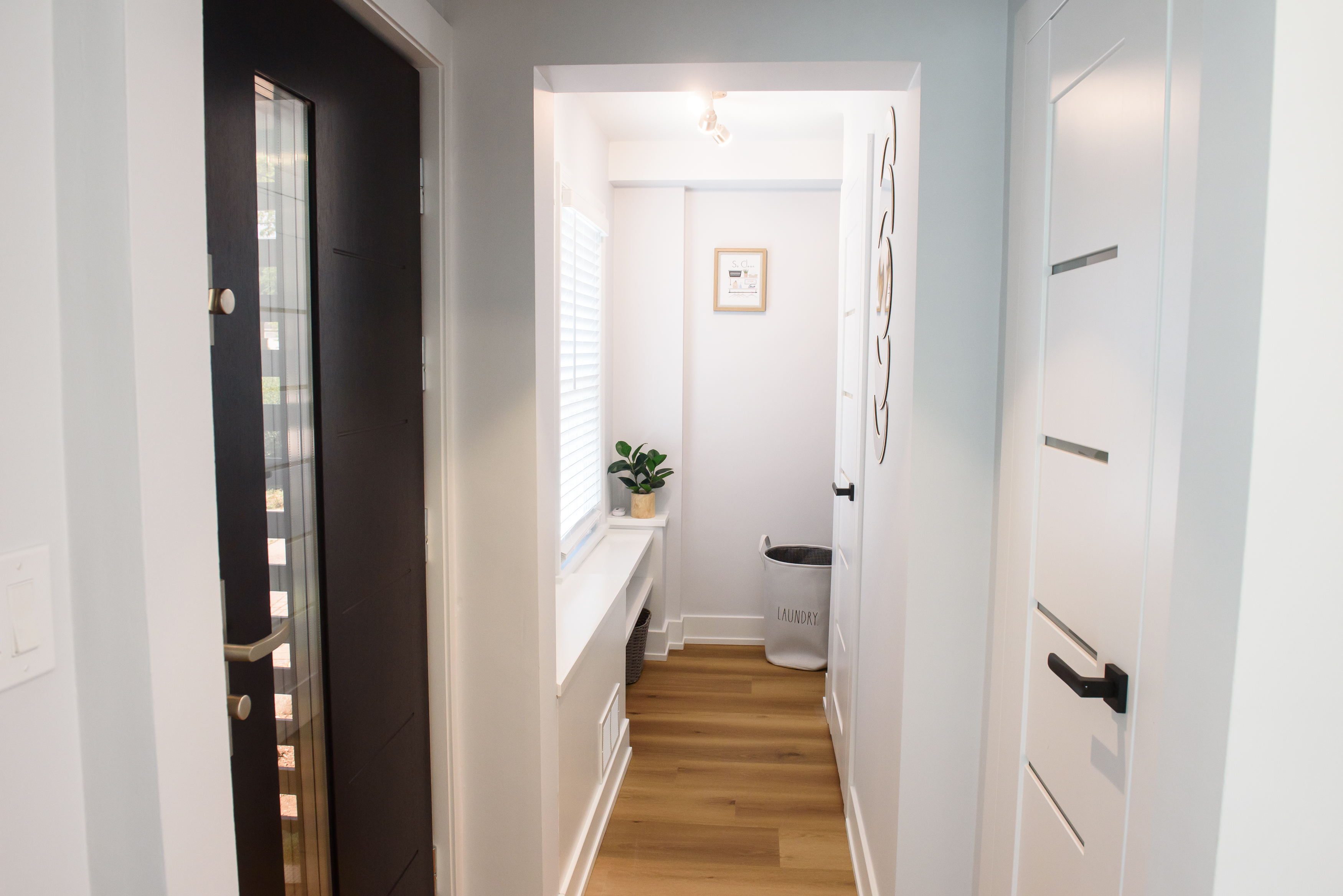 Narrow hallway with light wood floor, white walls, a dark entrance door on the left, a laundry basket at the end, and a small plant on a white shelf under a window.