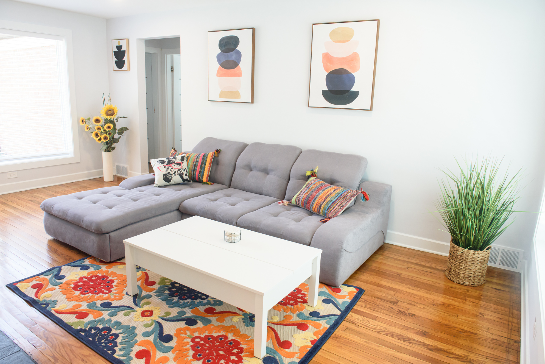 Bright living room with gray sectional sofa, colorful pillows, white coffee table on vibrant floral rug, and framed abstract art above.