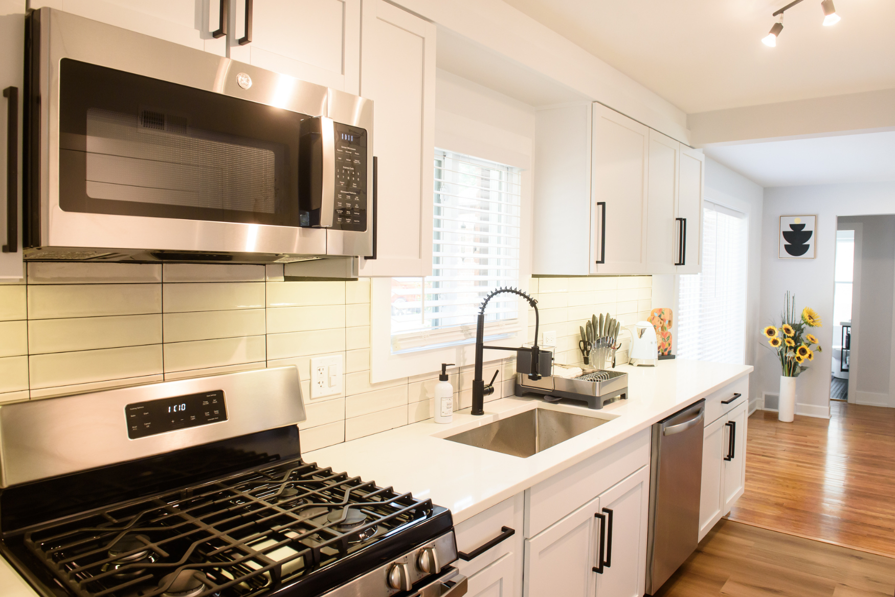 Modern kitchen with stainless steel stove, overhead microwave, white cabinets, black faucet, and a vase of sunflowers on wooden floor.