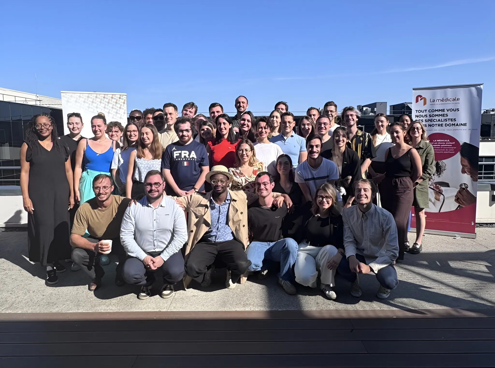 Groupe de personnes posant ensemble sur une terrasse ensoleillée avec des banderoles promotionnelles en arrière-plan.