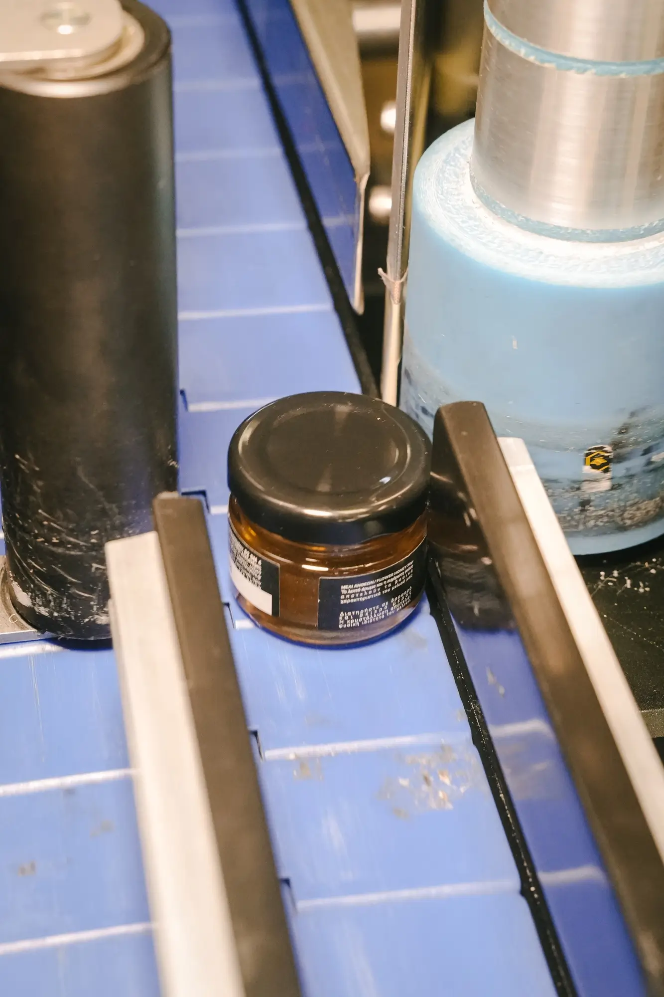 Small brown jar with a black lid moving on a blue conveyor belt between metal guides.