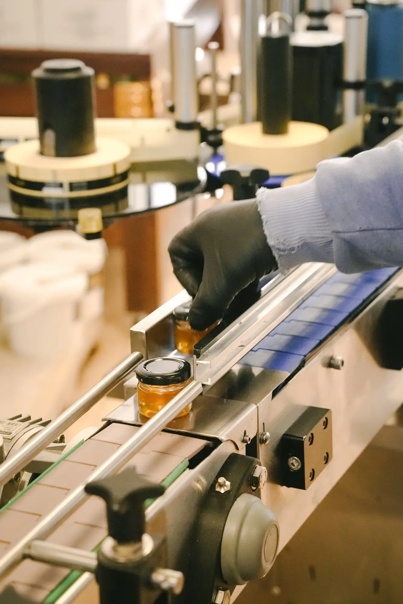 Gloved hand adjusting a small jar of honey on a conveyor belt in a production line.